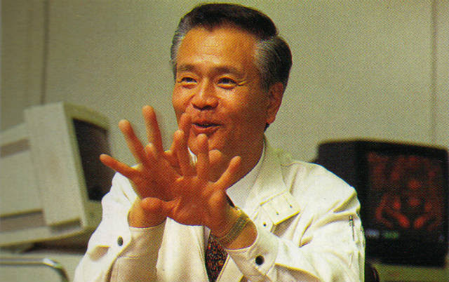 Retrato de Gunpei Yokoi hablando con las manos abiertas, delante de dos pantallas de ordenador